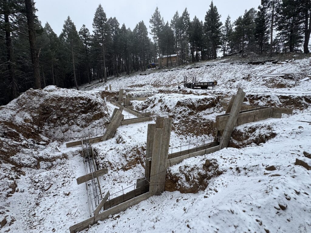Foundation forms set into a snow-covered, sloped mountain lot surrounded by pines